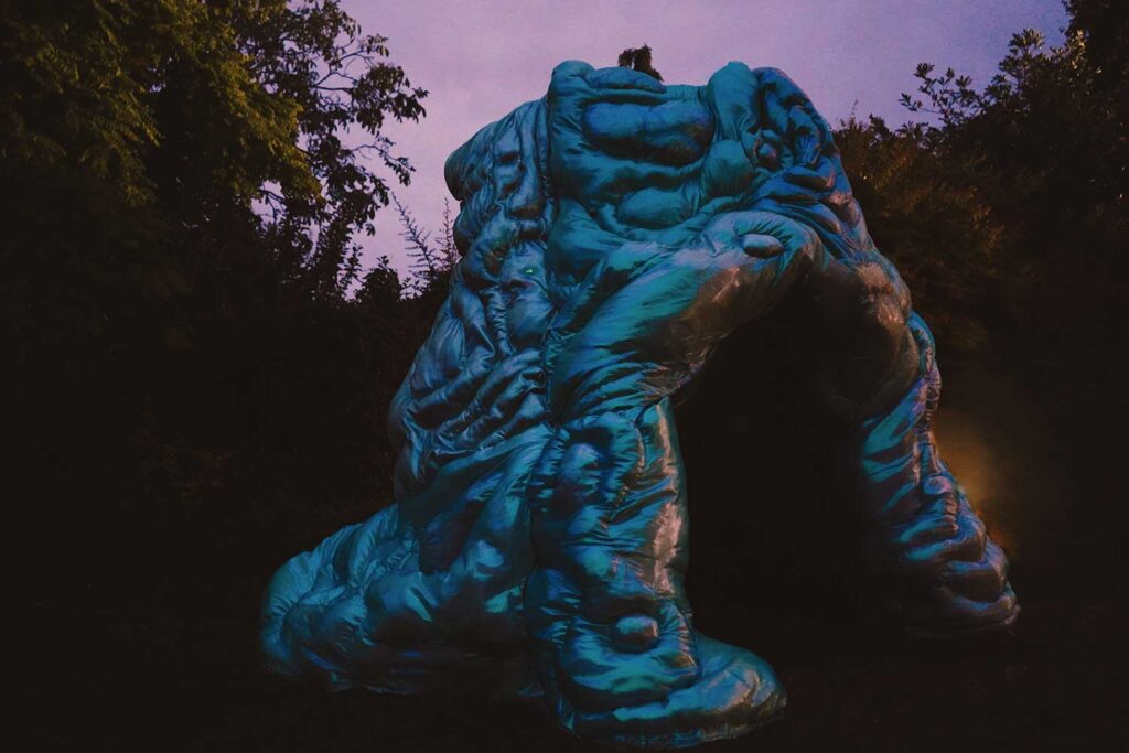 A large, abstract, blue sculptural structure stands outdoors at dusk, surrounded by dark trees and foliage.