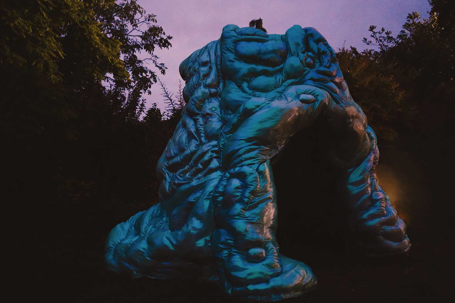 A large, abstract, blue sculptural structure stands outdoors at dusk, surrounded by dark trees and foliage.