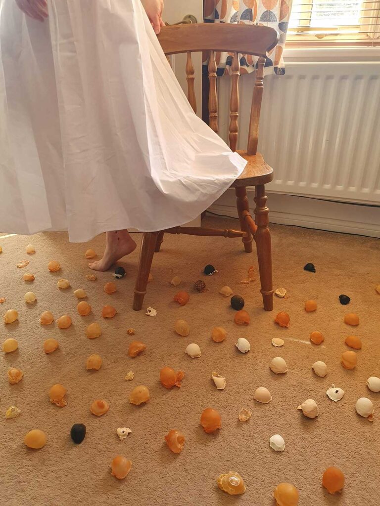 A person in a white dress stands on tiptoe beside a wooden chair, with numerous cracked eggs of various colorus scattered across a beige carpet in a sunlit room.