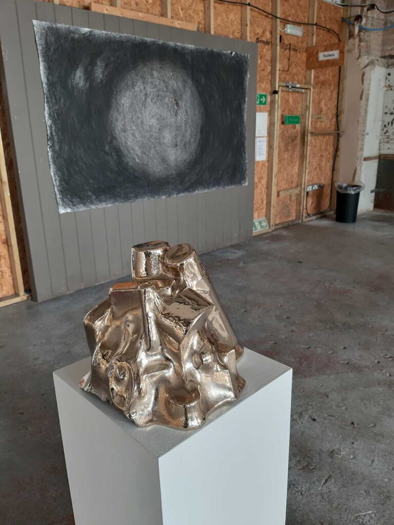 A metallic sculpture sits on a white pedestal in an industrial gallery space. A large, circular, textured black and white artwork is displayed on the wooden wall behind it.