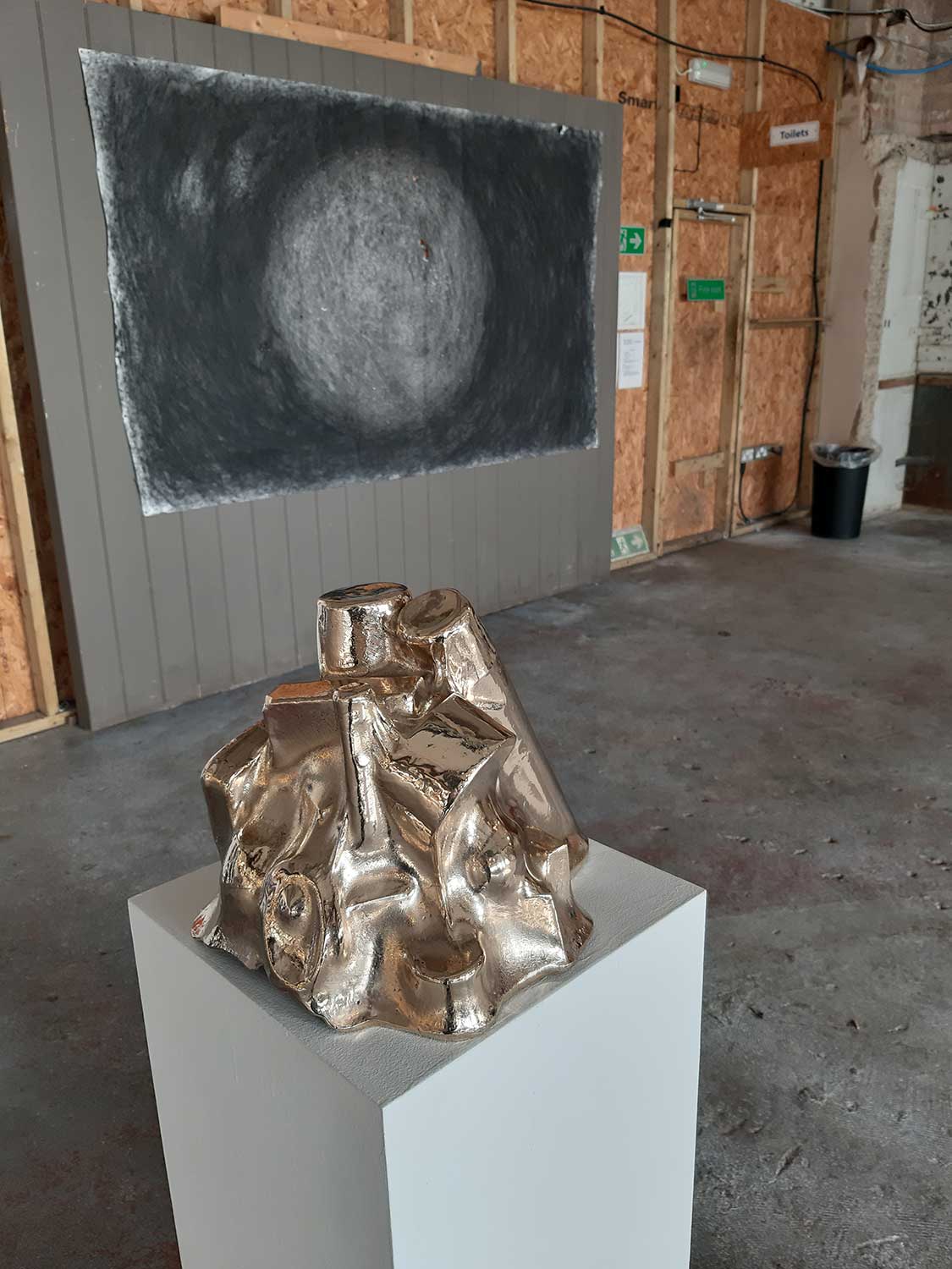 A metallic sculpture sits on a white pedestal in an industrial gallery space. A large, circular, textured black and white artwork is displayed on the wooden wall behind it.