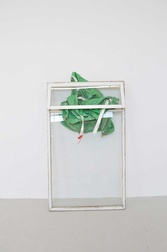 A white-framed glass window leans against a white wall. A green plastic bag is draped and tangled through the window frame, with some white straps hanging down. The scene is minimal and set in a bare room.