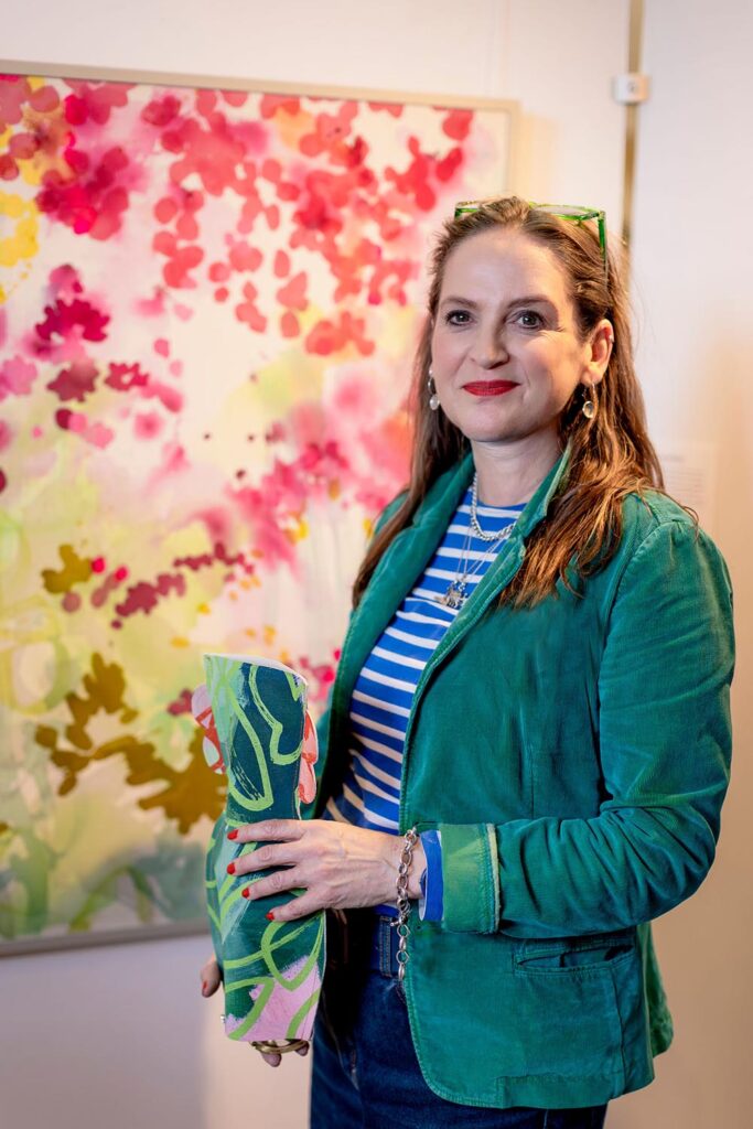 Lucy wears a green jacket and striped top. She stands in front of a colorful abstract painting, holding a patterned vase and smiling at the camera.