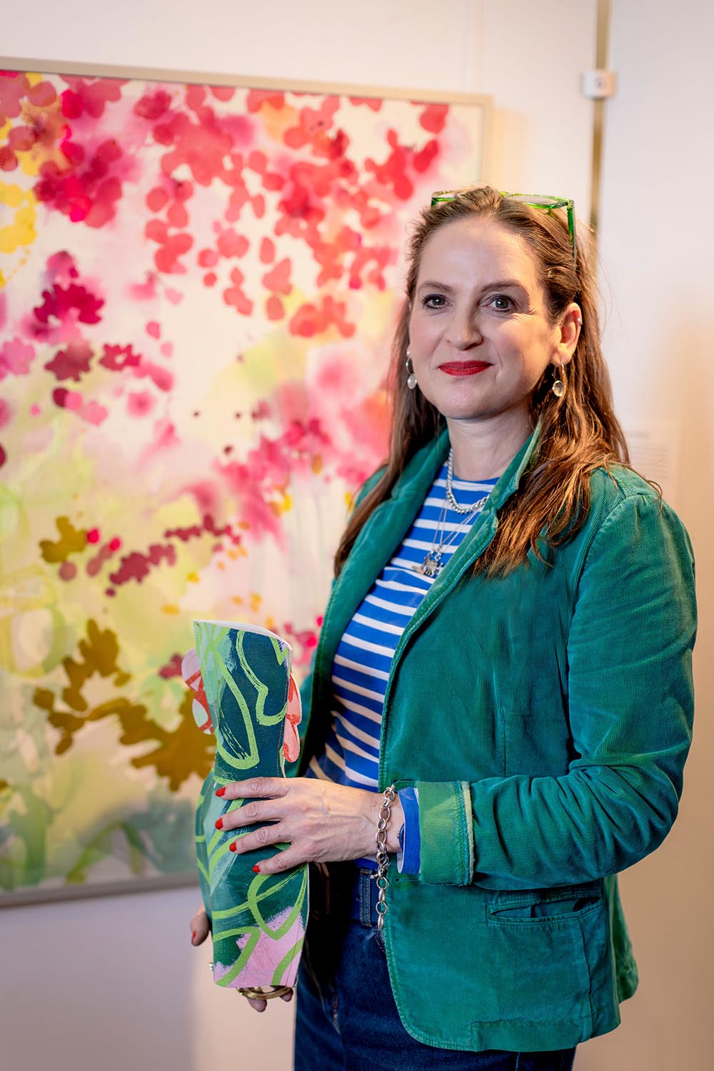 Lucy wears a green jacket and striped top. She stands in front of a colorful abstract painting, holding a patterned vase and smiling at the camera.