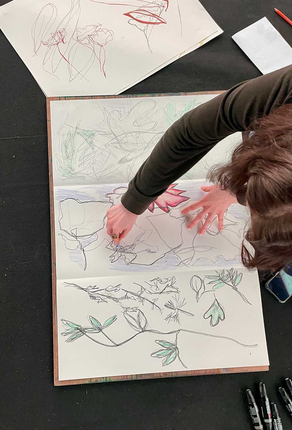 A person with brown hair sketches floral designs, surrounded by drawing supplies and paper. The person's arm is extended over the page.