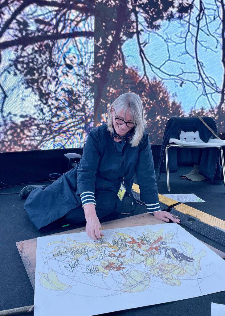 A person kneels on the floor, drawing on a large sheet of paper filled with colorful sketches. Behind her is a large screen displaying tree branches.