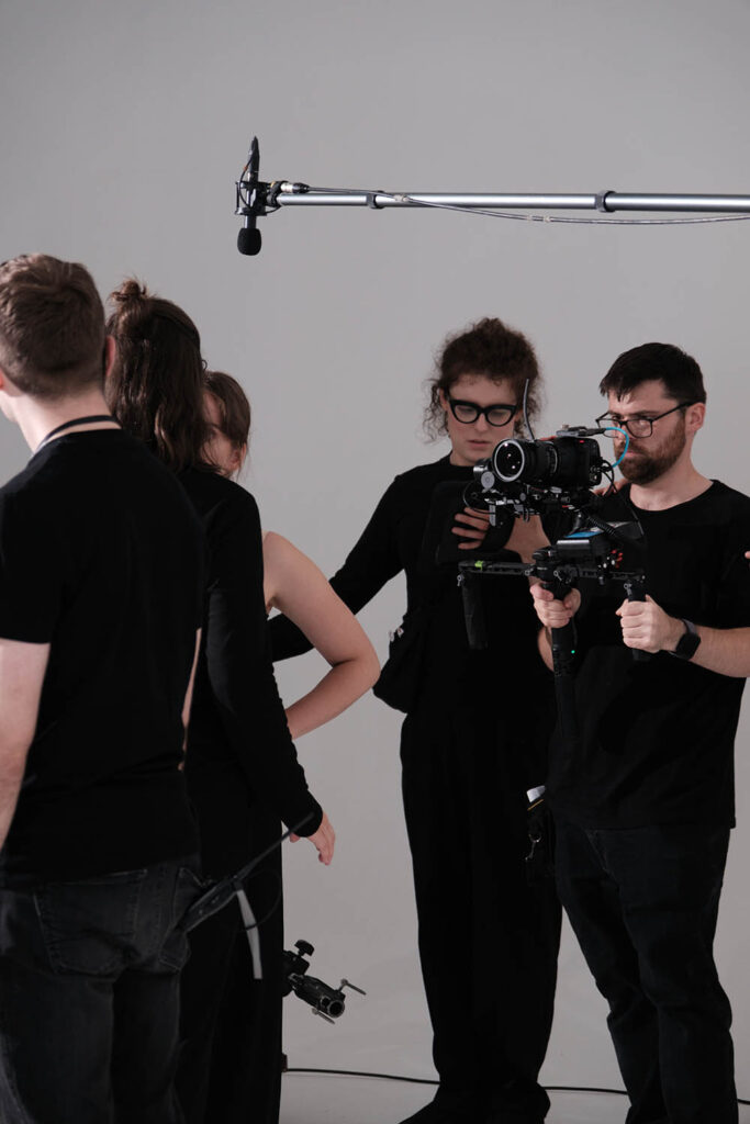 A film crew, dressed in black, works in a studio with a camera and boom microphone. Two crew members handle the camera while others stand nearby.