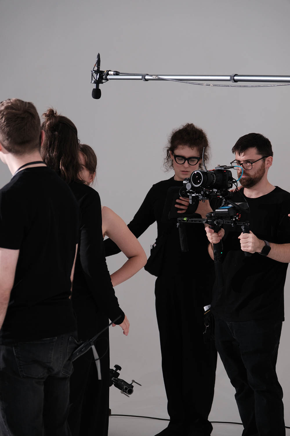 A film crew, dressed in black, works in a studio with a camera and boom microphone. Two crew members handle the camera while others stand nearby.