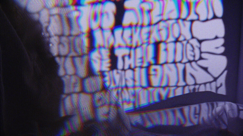 Blurry, distorted letters in a bold, rounded font are projected onto a surface, creating a colorful, psychedelic effect with purple hues. The text is mostly unreadable due to the distortion.