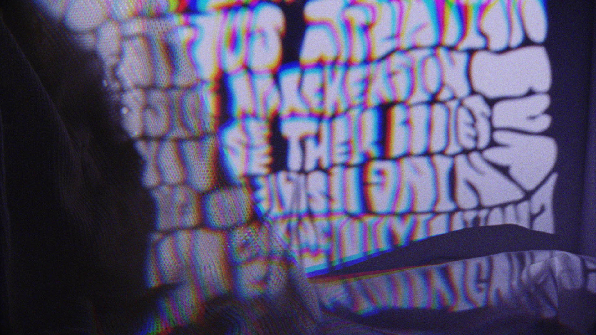 Blurry, distorted letters in a bold, rounded font are projected onto a surface, creating a colorful, psychedelic effect with purple hues. The text is mostly unreadable due to the distortion.