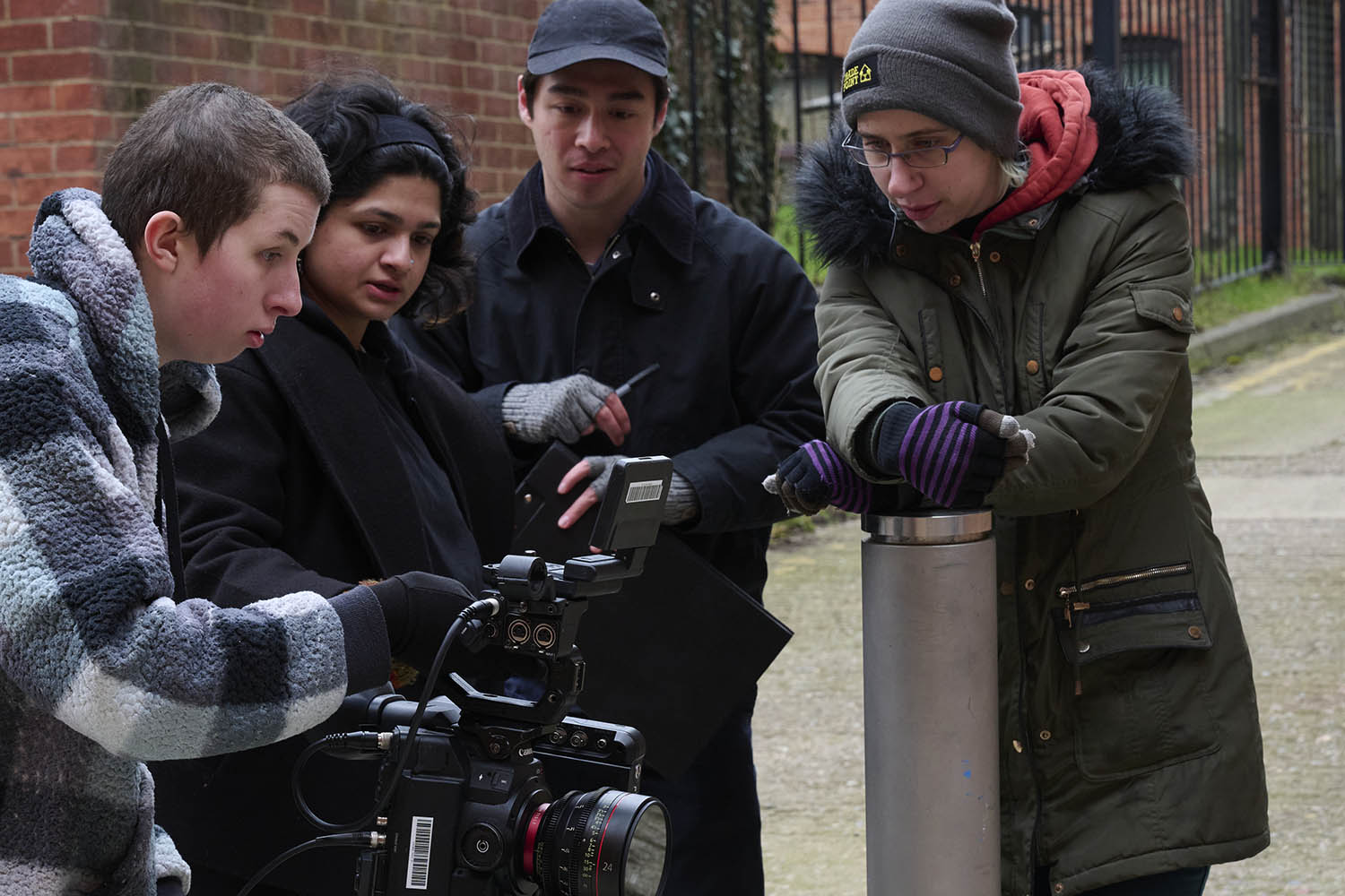 Four people are gathered outdoors around a professional video camera, focusing intently. One person adjusts the camera while the others watch. They are dressed warmly in jackets and hats.