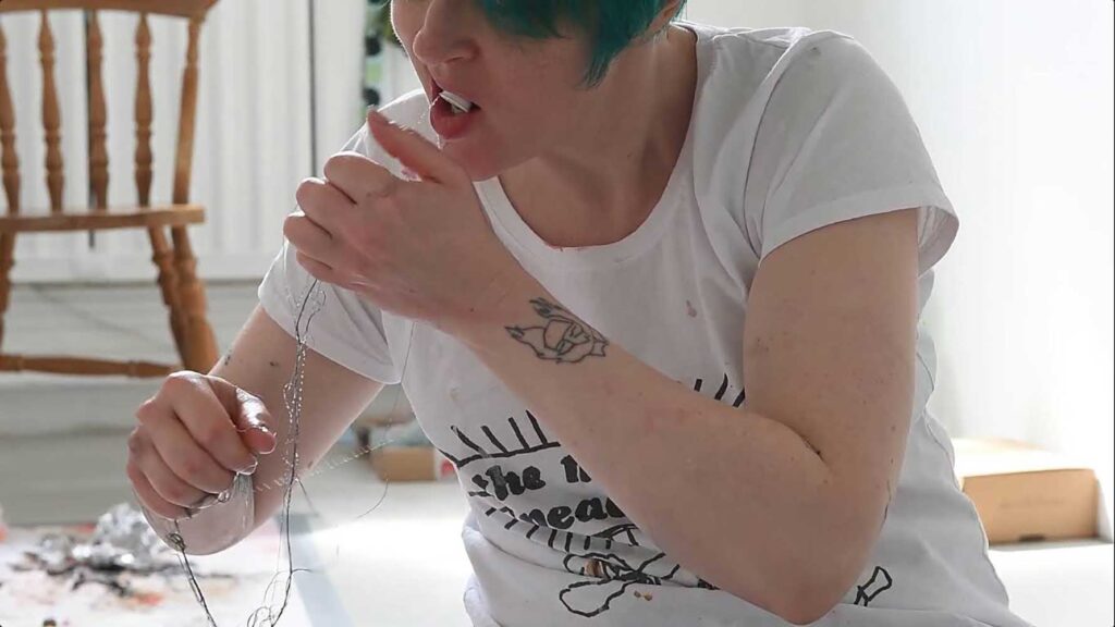 A person with short teal hair holds a piece of wire up to their mouth while seated indoors. They wear a white t-shirt with black text and a graphic design. A wooden chair is visible in the background.