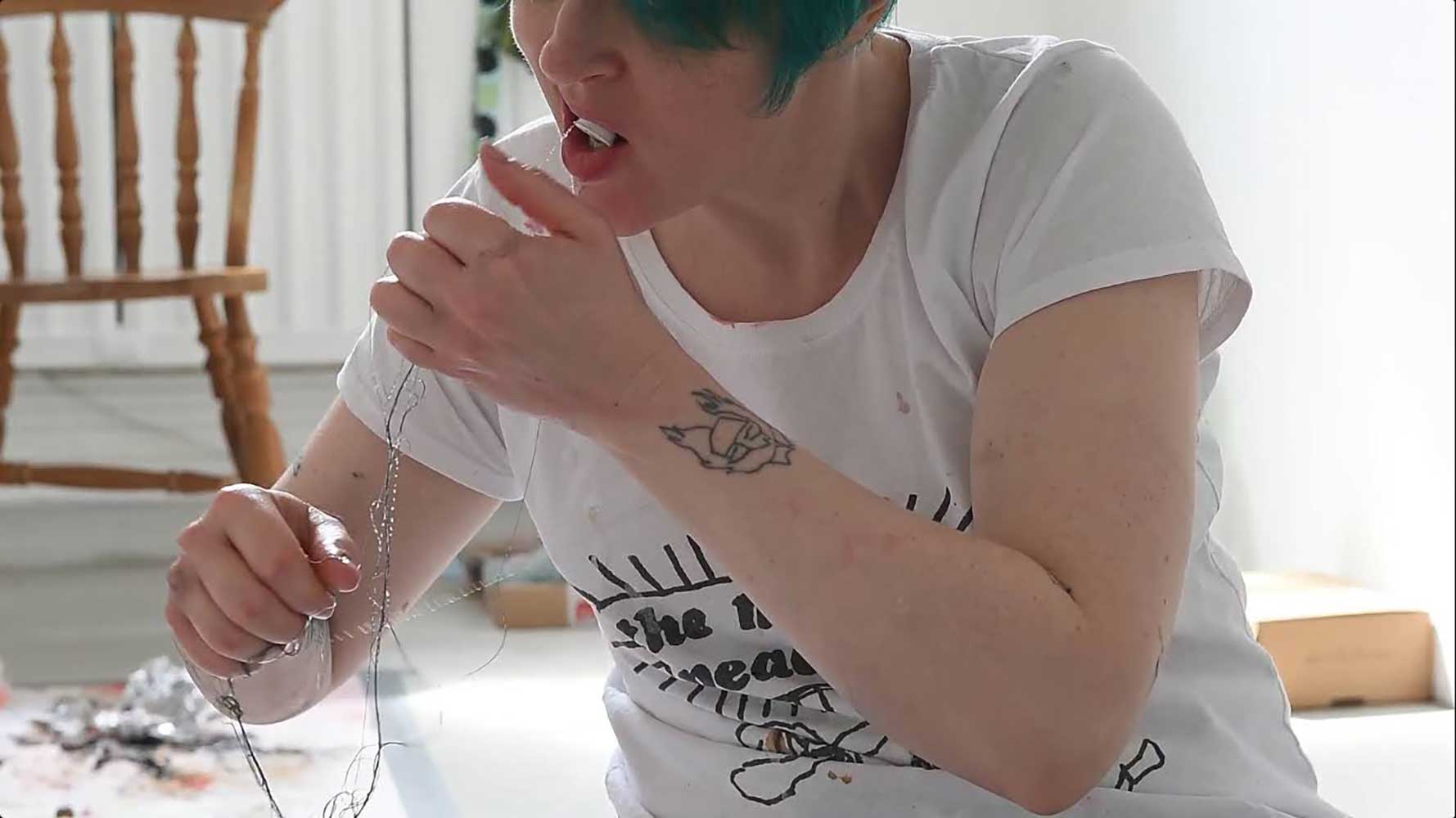 A person with short teal hair holds a piece of wire up to their mouth while seated indoors. They wear a white t-shirt with black text and a graphic design. A wooden chair is visible in the background.