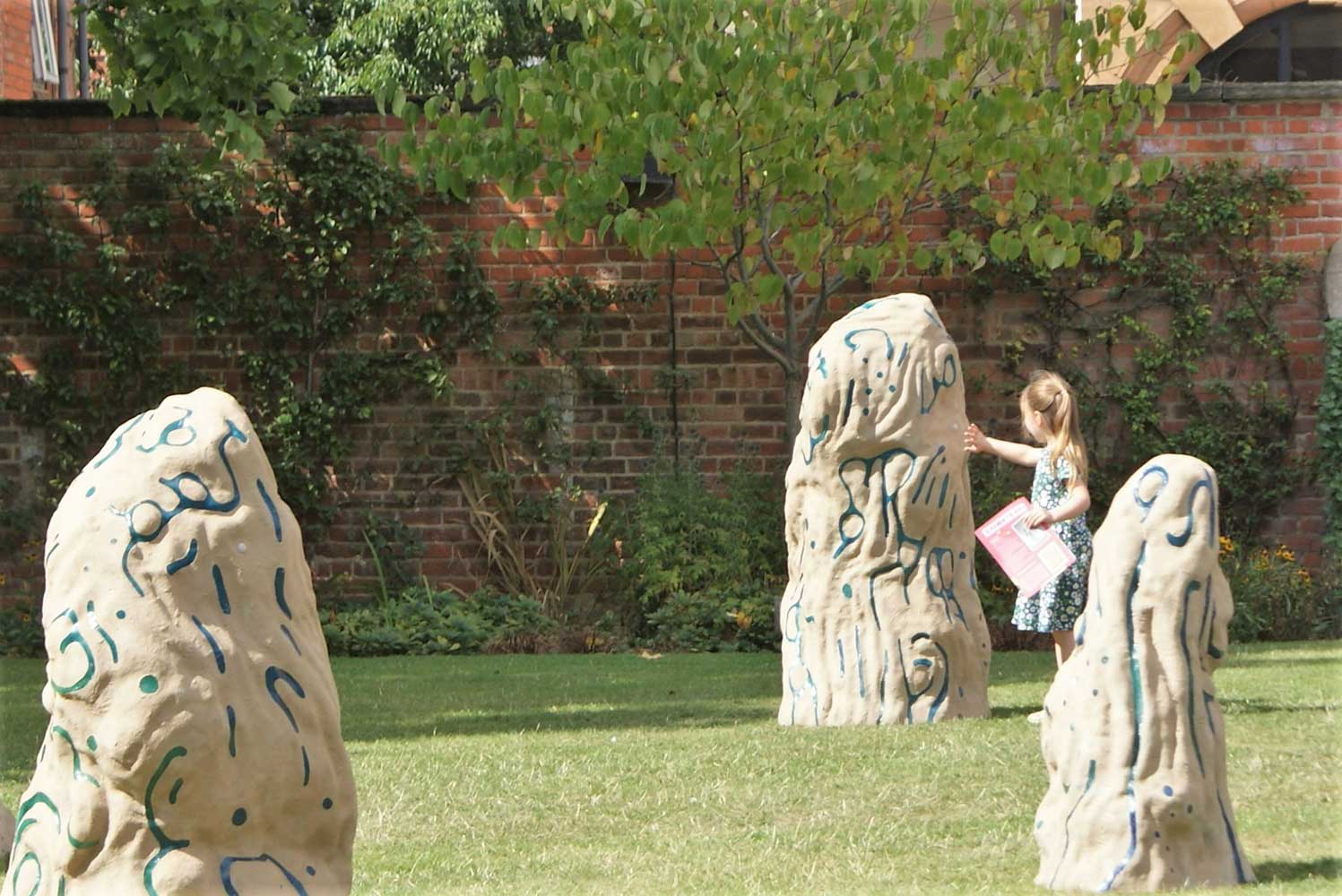A child in a summer dress stands near a tall, abstract stone sculpture in a grassy garden. More similar sculptures and a brick wall with greenery are in the background.