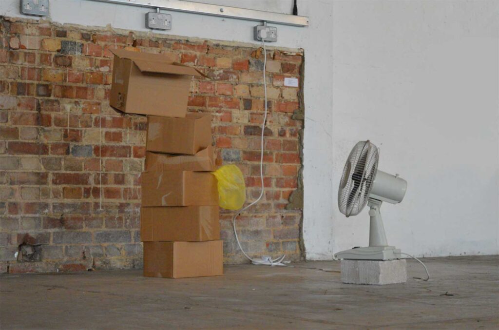 A small electric fan on a concrete block faces a stack of precariously balanced cardboard boxes, with a yellow plastic bag caught between them, set against a brick and white wall.