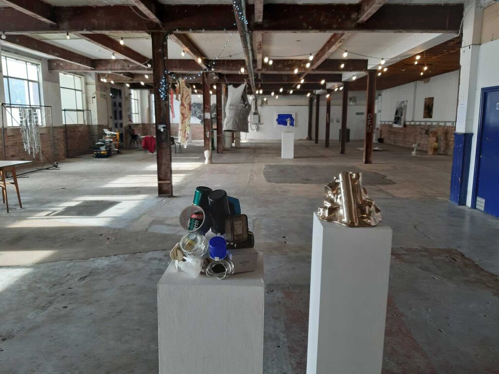 A spacious art gallery with exposed beams and brick, featuring sculptures on white pedestals, industrial lighting, and various artworks displayed along the walls.