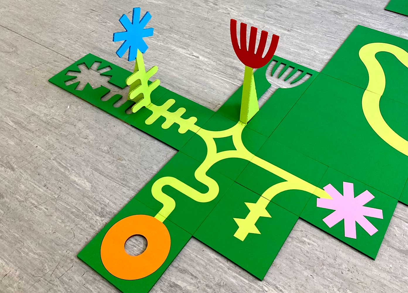 Colourful, abstract shapes are arranged on interlocking green tiles placed on a grey floor, creating a playful design.