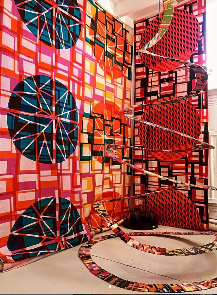 Colourful, geometric-patterned fabric hangs on walls and forms looping, ribbon-like spirals in a room. Dominant colors include red, pink, orange, and blue with bold circles and lines.