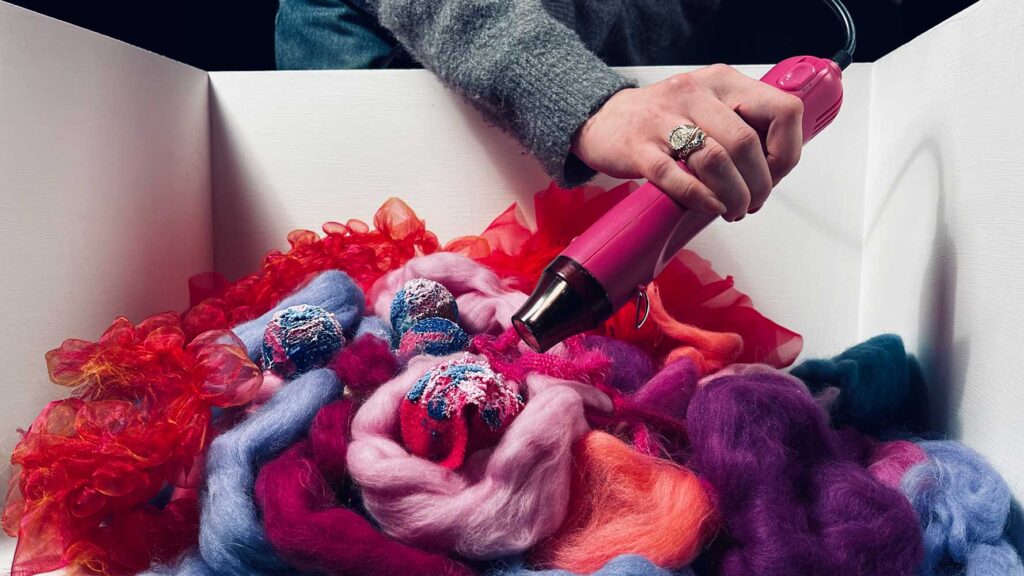 A person uses a pink heat gun over colourful wool fibers and yarn, with red, purple, orange and pink textures arranged inside a white box.