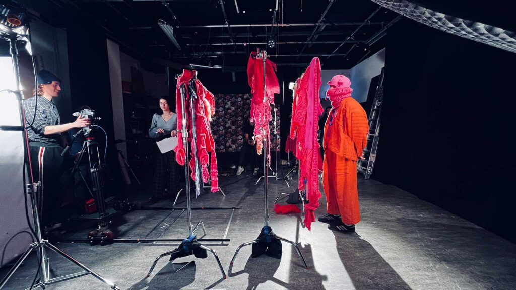 A studio setting with people filming a person in orange clothing and a pink mask, standing near stands holding bright pink fabrics. Cameras, lighting equipment and cables are visible throughout the scene.