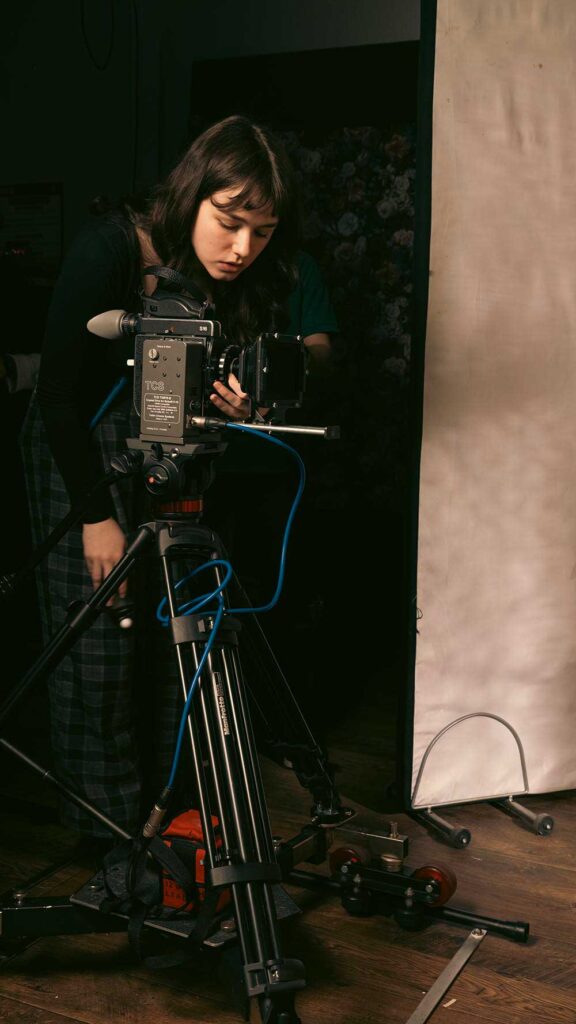 A person adjusts a professional video camera mounted on a tripod in a studio setting, with cables attached to the camera and lighting equipment visible nearby.