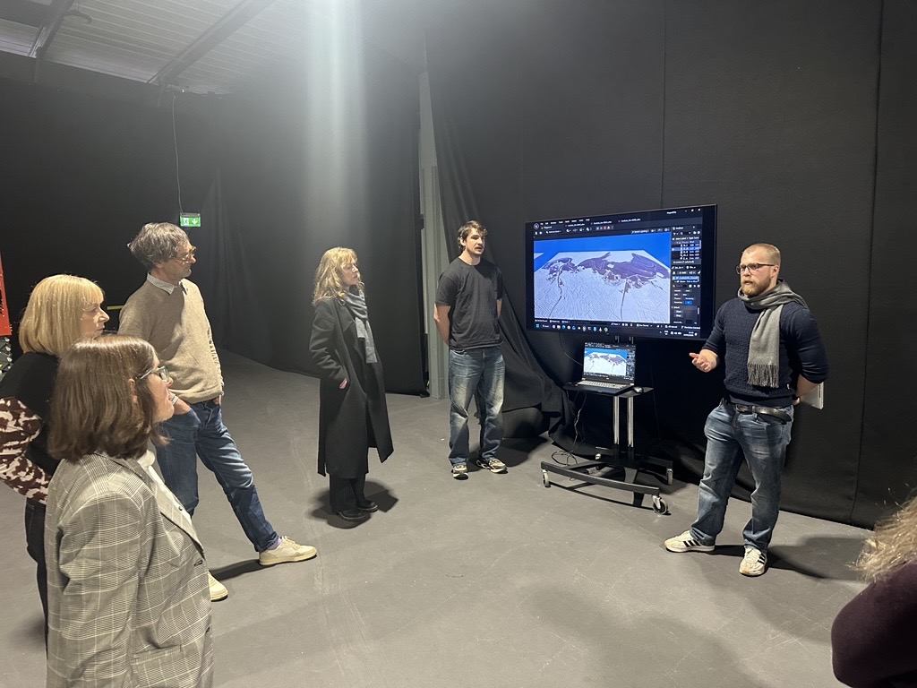 A group of people stands in a room with dark walls, listening to a man giving a presentation in front of a large screen displaying a mountain landscape image. A laptop is connected to the screen on a wheeled stand.