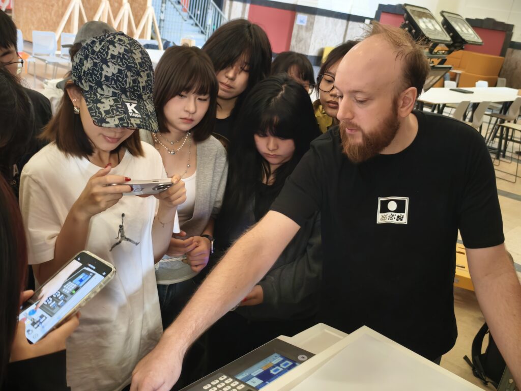 A bearded man demonstrates how to use a machine to a group of attentive students, some of whom are taking photos or videos with their phones.