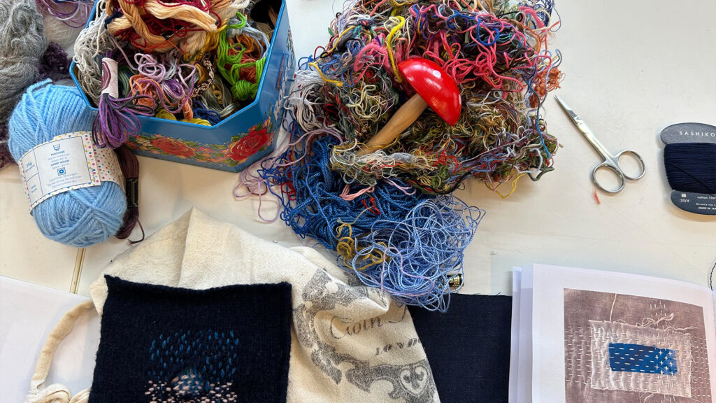 A tabletop with tangled colored threads, a red-handled darning tool, scissors, blue yarn, a floral tin, embroidery projects, and an open book showing a stitched blue patch.