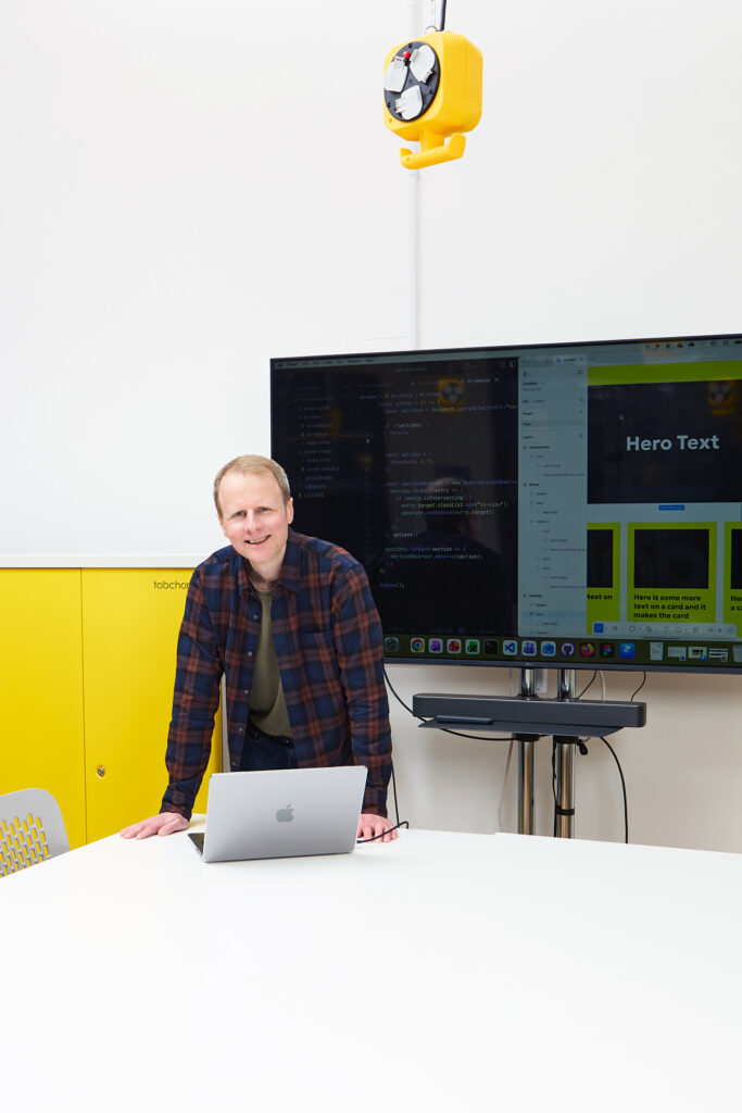 A person in a plaid shirt stands behind a white table with a laptop, in front of a large screen displaying code and a website layout, in a modern, bright office with yellow accents.
