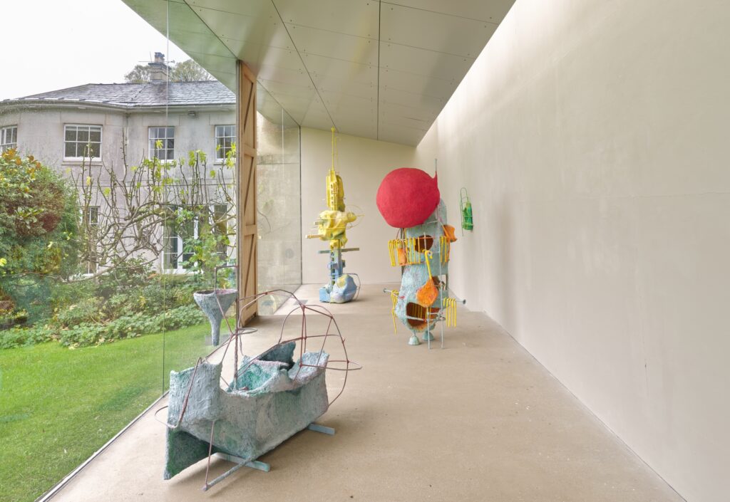 Colorful abstract sculptures are displayed in a bright gallery space with large glass walls overlooking a green garden and an old house. The sculptures are made of mixed materials and irregular shapes.