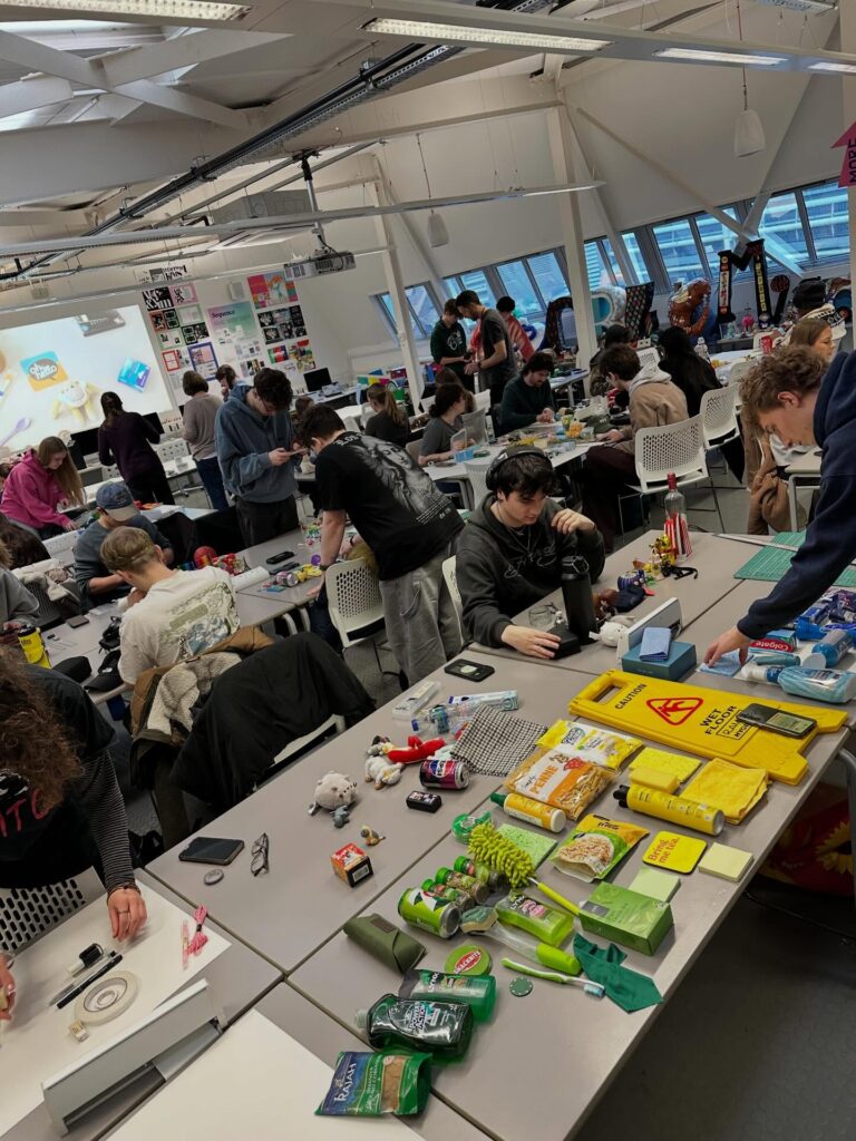 A busy classroom filled with students working on creative projects at tables covered with various craft materials, tools, and colorful objects. Some students are seated while others stand and collaborate.