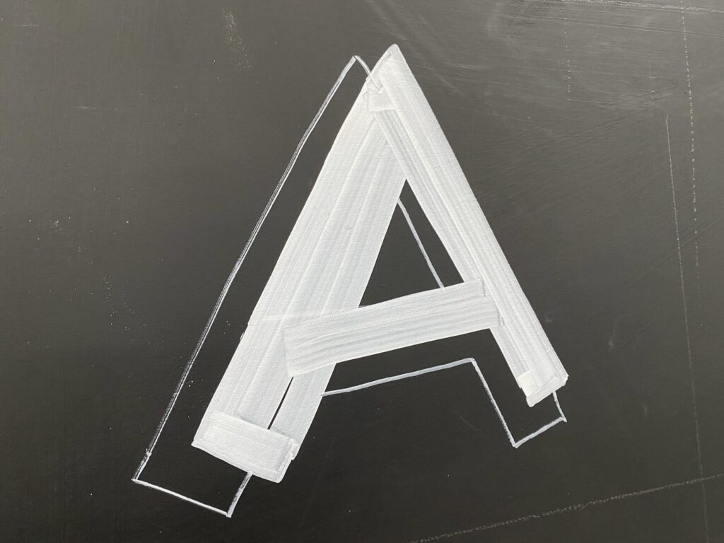 A large, white, uppercase letter "A" is drawn on a blackboard in thick, bold strokes, with the outline of the letter visible in thin white lines.
