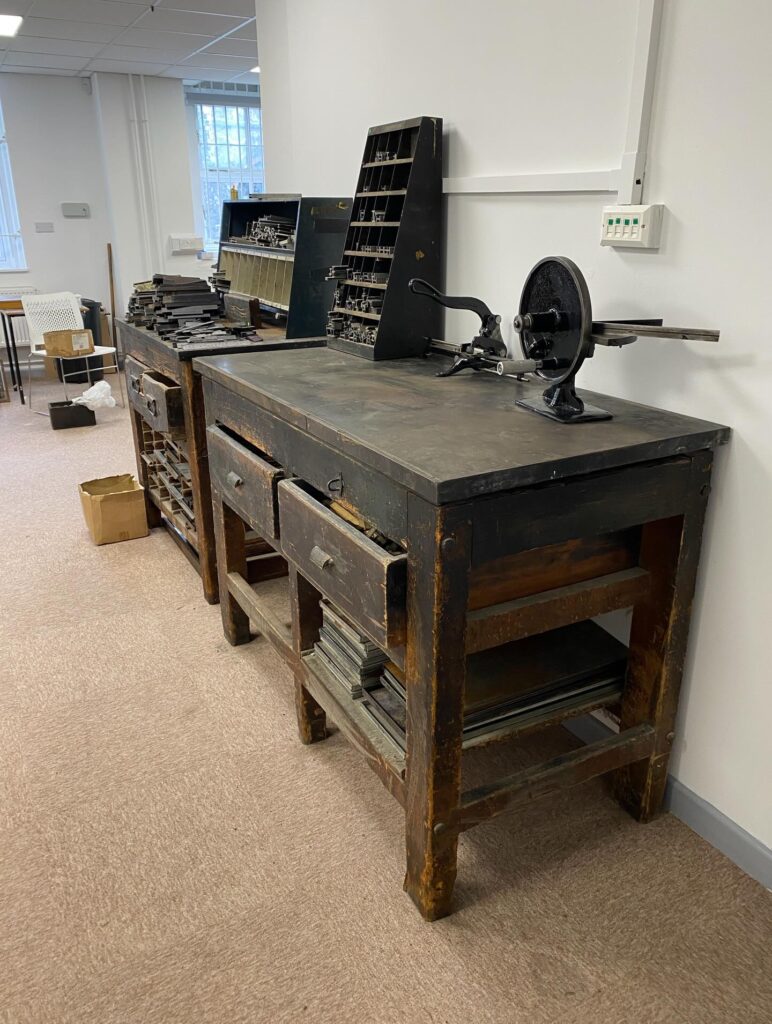 A vintage wooden workbench with drawers stands against a wall, holding old typesetting equipment and a small printing press. Other trays and type cases are arranged nearby, in a well-lit room with beige carpet.