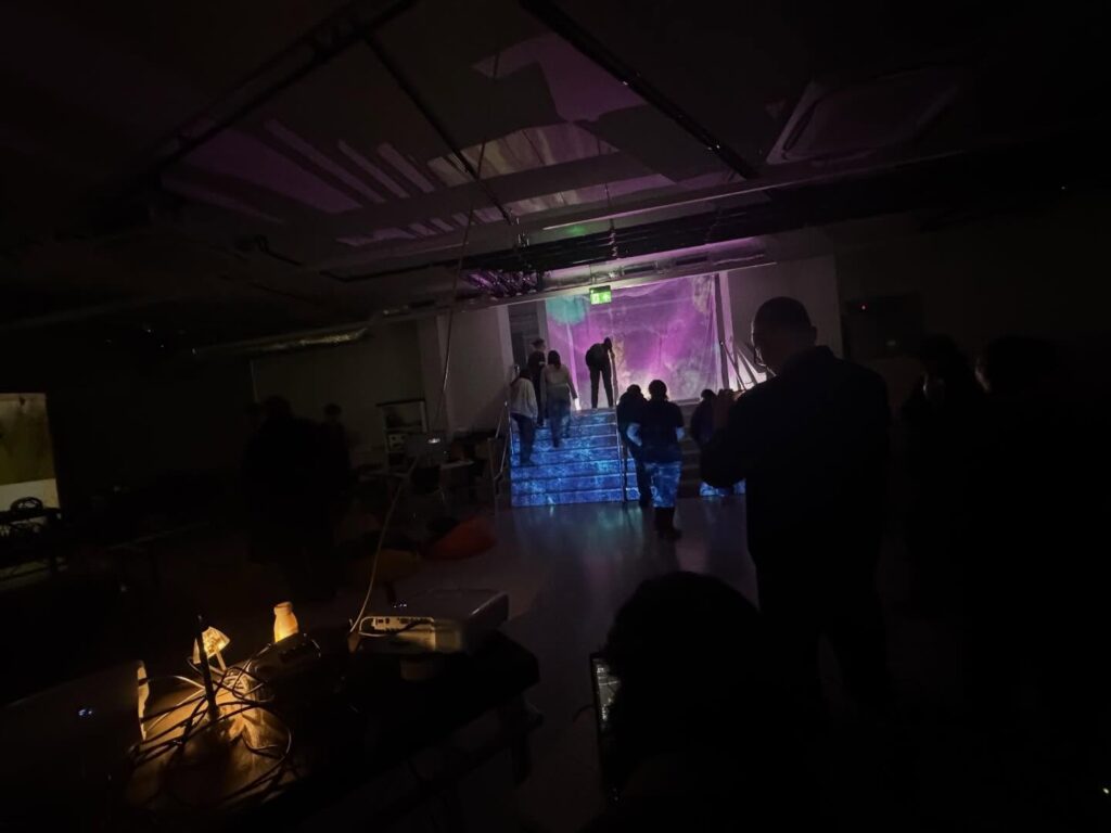 A dimly lit room with people gathered, some standing on illuminated steps leading to a glowing purple backdrop; a table with lights and equipment is visible in the foreground.