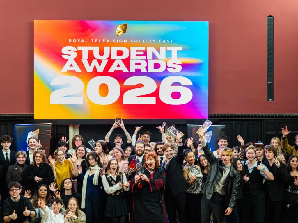A large group of students celebrate and pose with trophies in front of a colorful screen reading "Royal Television Society East Student Awards 2026." Many are smiling, cheering, and raising their hands.