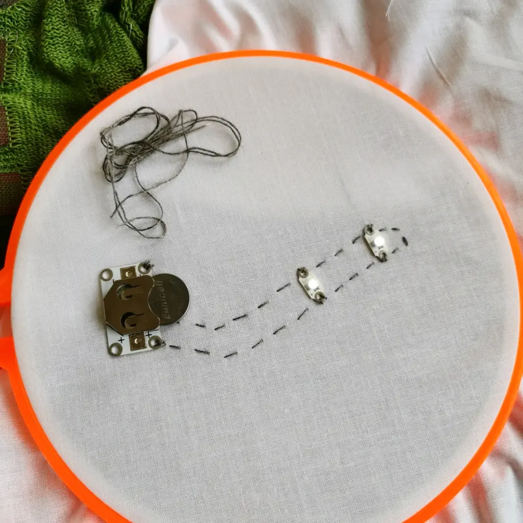 An embroidery hoop with white fabric shows a few stitched black lines, electronic components sewn on, and a bundle of black thread resting on the fabric. A green textured fabric is partially visible in the background.