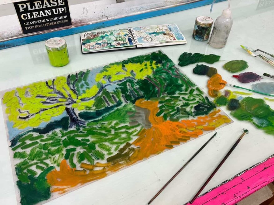 A colorful landscape painting of a tree and path is in progress on a table, surrounded by paintbrushes, paint, and art supplies. A sign reading “PLEASE CLEAN UP!” is visible in the background.