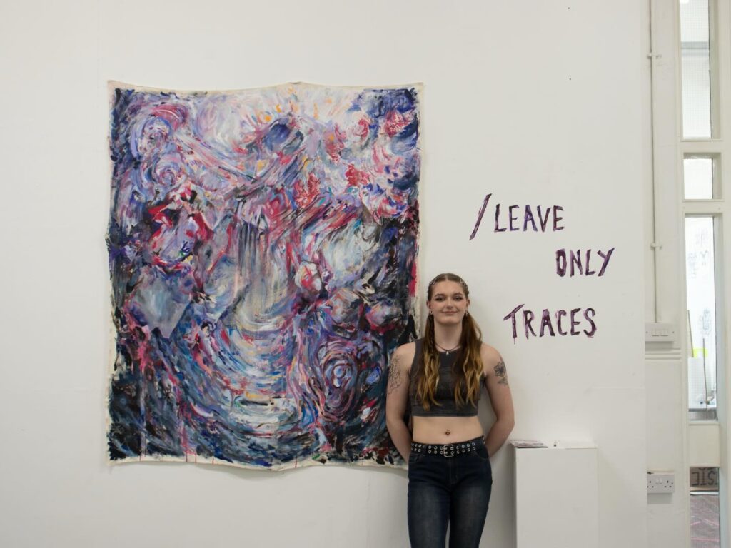 A person with long hair and tattoos stands smiling next to a colorful, abstract painting. On the wall beside them, purple text reads "/ LEAVE ONLY Traces." The setting appears to be an art gallery or exhibition space.