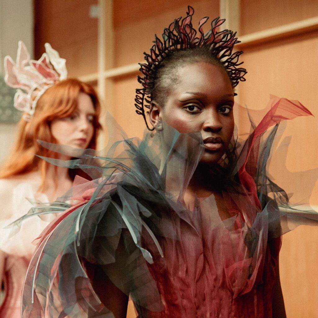 Two models wearing high-fashion tulle dresses; the model in front wears a dramatic burgundy and black gown with spiked tulle details, while the model behind has red hair and a light pink dress with a delicate headpiece.