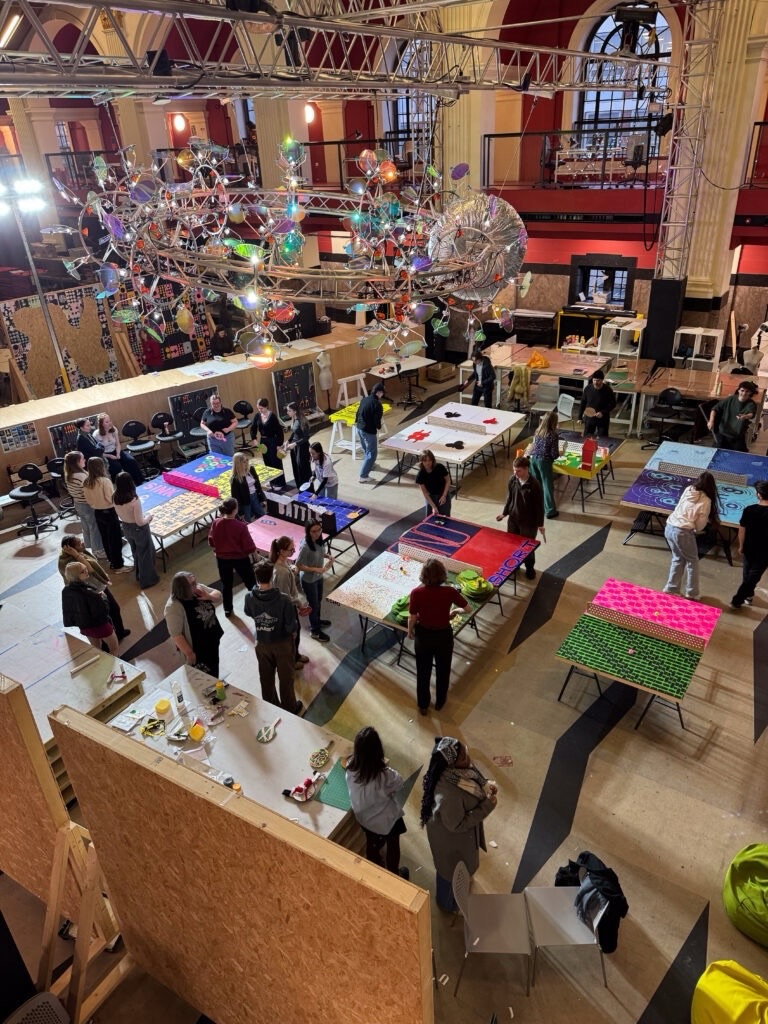 A large, brightly lit hall with high ceilings and chandeliers, where people are gathered around tables covered in colorful art projects, working and socializing in a creative, collaborative space.