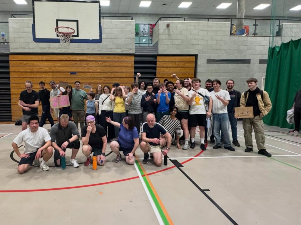 A large group of people, some in sportswear, pose together in a gymnasium with a basketball hoop in the background. Some are kneeling, while others stand in the back, smiling and cheering.