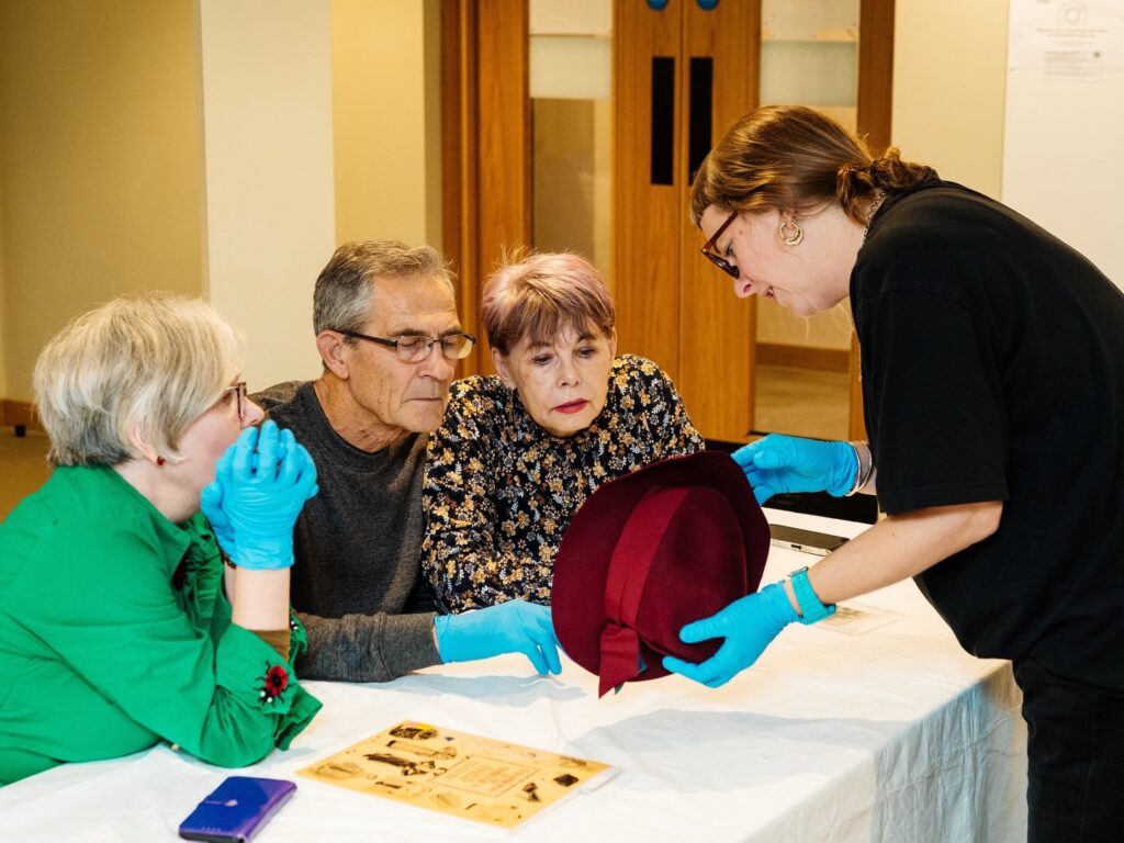 Three older adults examine a vintage red hat held by a woman wearing gloves, while sitting at a table with documents and a phone. All participants are focused, wearing blue gloves, in a well-lit indoor setting.