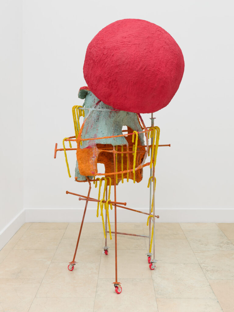 A colorful abstract sculpture with a large red sphere on top of a textured turquoise and orange structure, supported by yellow and orange metal rods on wheels, set against a plain white wall and tiled floor.