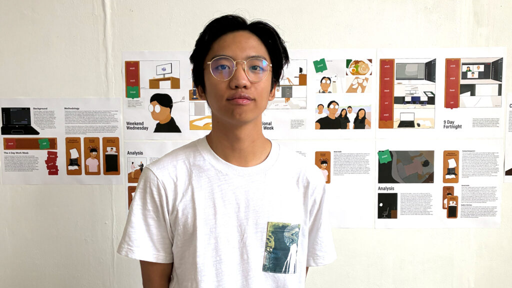A person with glasses and short dark hair, wearing a white t-shirt with a graphic pocket, stands in front of a wall displaying several colorful infographics and illustrated posters.