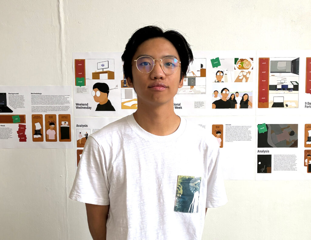 A young person with glasses and a white t-shirt stands in front of a wall displaying colorful graphic illustrations and charts. The background appears to be an exhibition or project presentation.