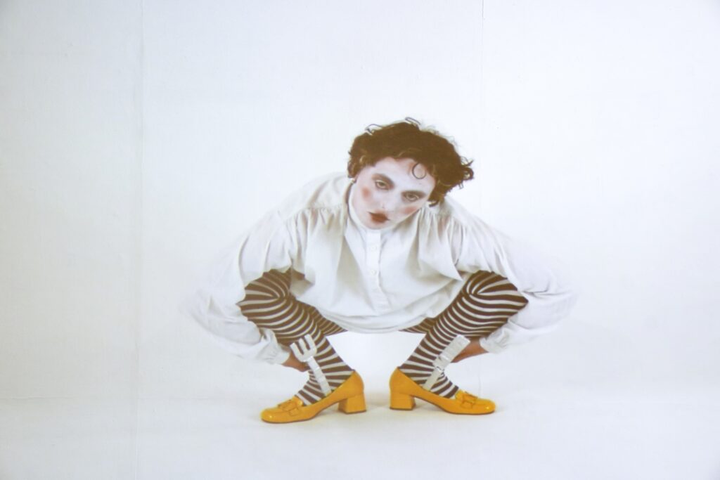 A person with white face makeup, red lips, and curly dark hair squats against a white background wearing a loose white shirt, black and white striped tights, and bright orange heels.