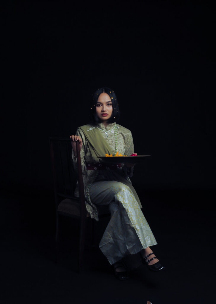 A woman wearing a traditional outfit sits on a chair against a dark background, holding a tray with colourful flowers. She has a calm expression, and small decorative ornaments are in her hair.