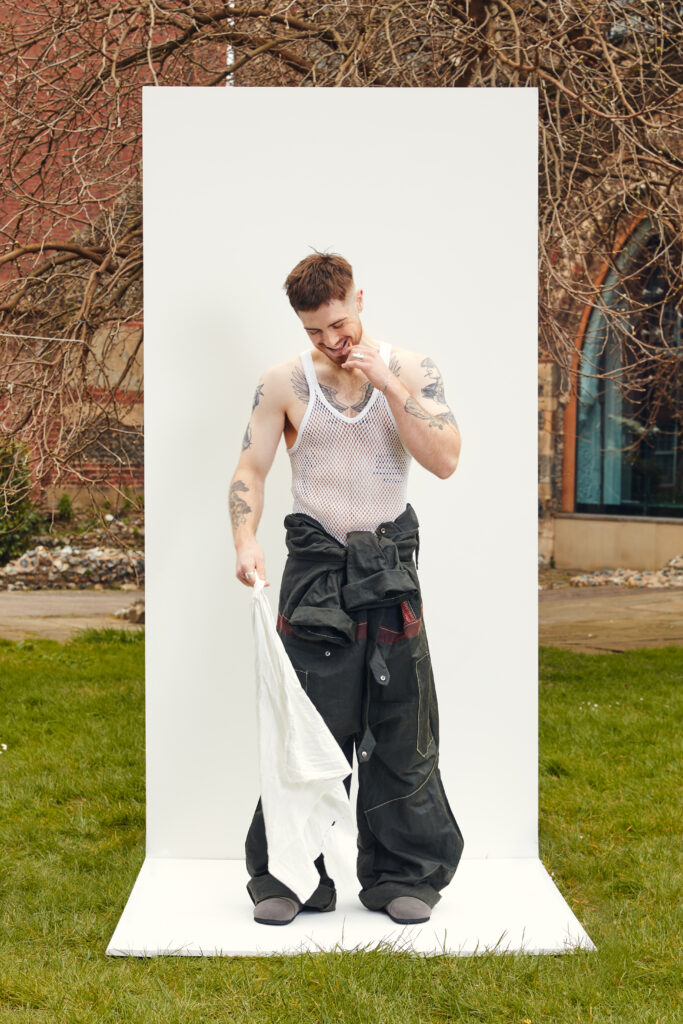 A man with tattoos wearing a white mesh tank top and baggy black pants stands on a white platform outdoors, holding a white cloth and smiling, with bare trees and a building in the background.