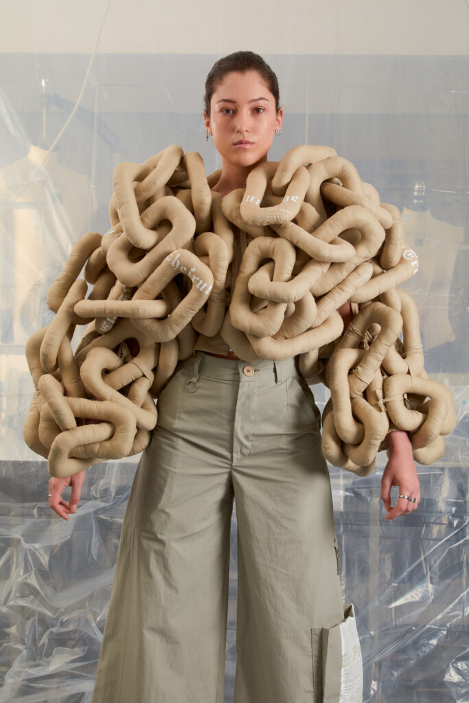 A person stands facing forward, wearing a unique beige top made of large, padded, interlocking chain-like loops. They also wear wide-leg light gray pants. The background features clear plastic sheets and soft lighting.