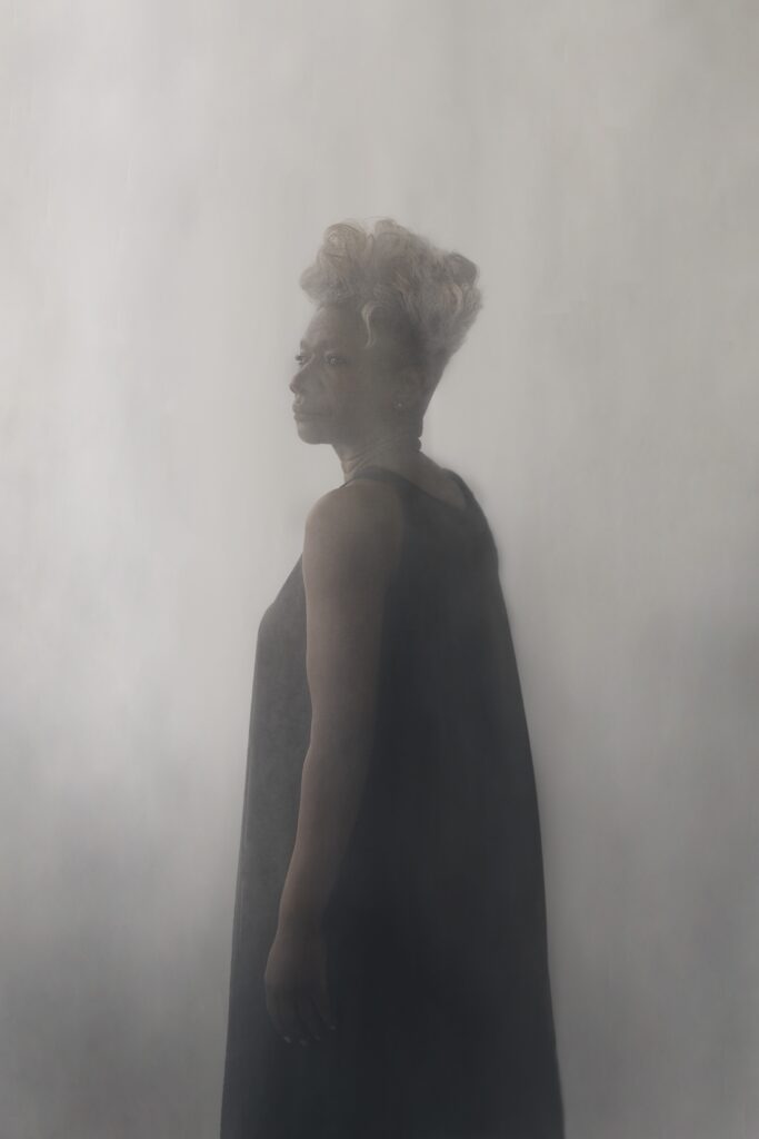 A person with short, curly white hair stands in profile, wearing a sleeveless black dress against a soft, misty, light gray background. The lighting creates a dreamy, ethereal atmosphere.